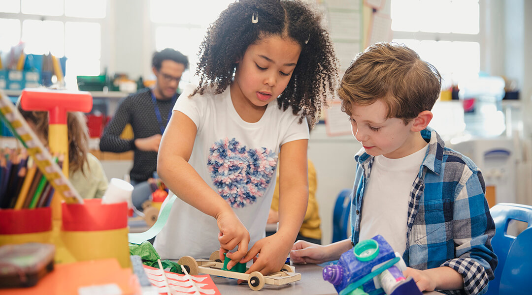 Elementary school children build 3D models using recycled materials during class.