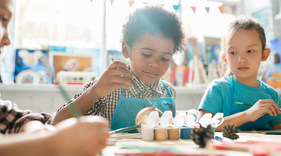 Serious child, about five, and his classmate sitting by desk and painting decorations.