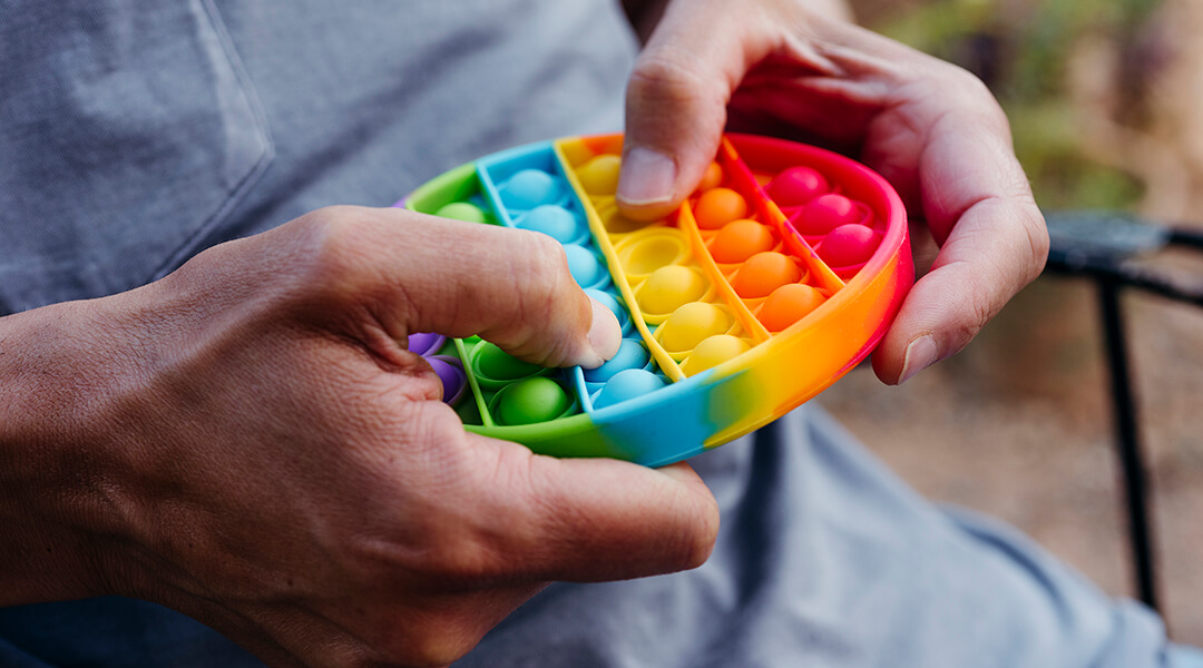 Close cropped image of an adult sitting outdoors, popping bubbles of a destressing toy patterned with the colors of the rainbow.