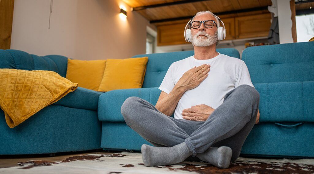 Mature senior adult sitting on floor of living room practicing guided meditation at home, with eyes closed and headphones on.