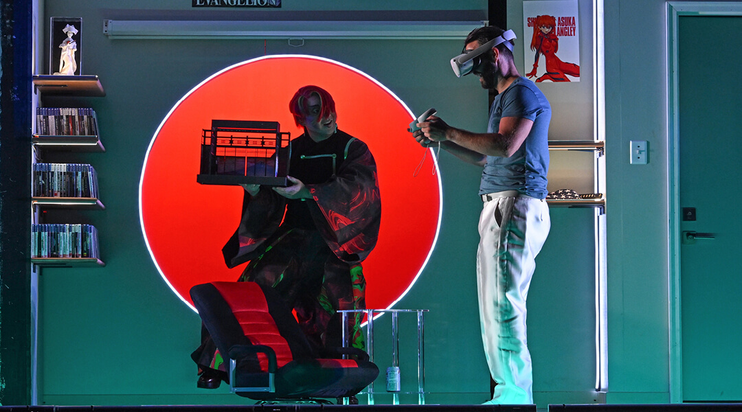 An actor wearing a VR headset on stage during a play, near a character obscured by dark clothing.